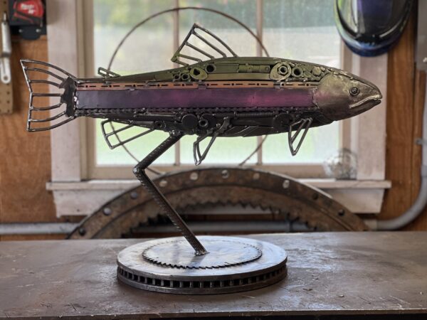 Scrap metal rainbow trout sculpture mounted on a gear-based stand, displayed in a workshop with a window in the background
